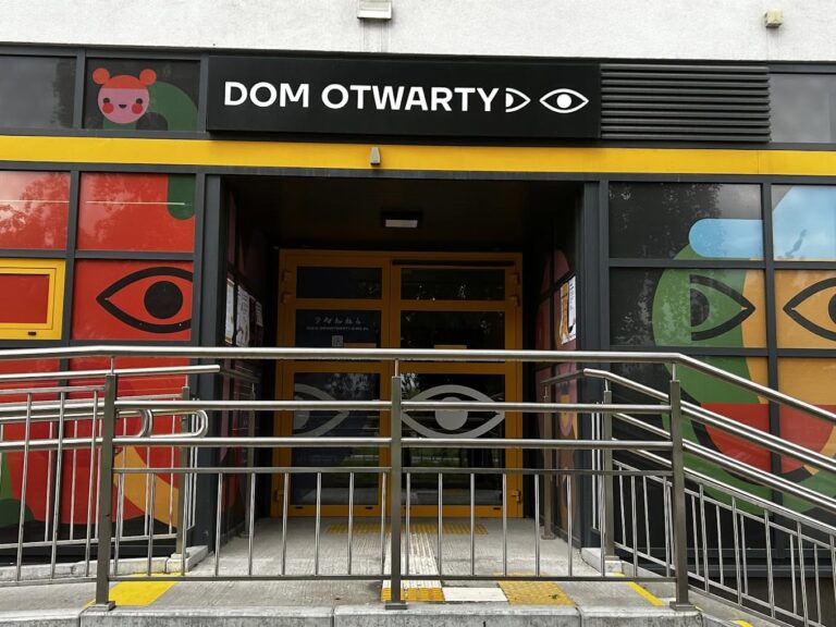 Wejście do budynku "Dom Otwarty" z pomarańczowymi drzwiami, metalową poręczą i podjazdem dla wózków, oznaczonym żółtymi pasami faktury ostrzegawczej. Nad wejściem czarny szyld z białym napisem "DOM OTWARTY" i symbolem oka, a witryny po bokach zdobione kolorowymi ilustracjami z motywem oczu i postaci.