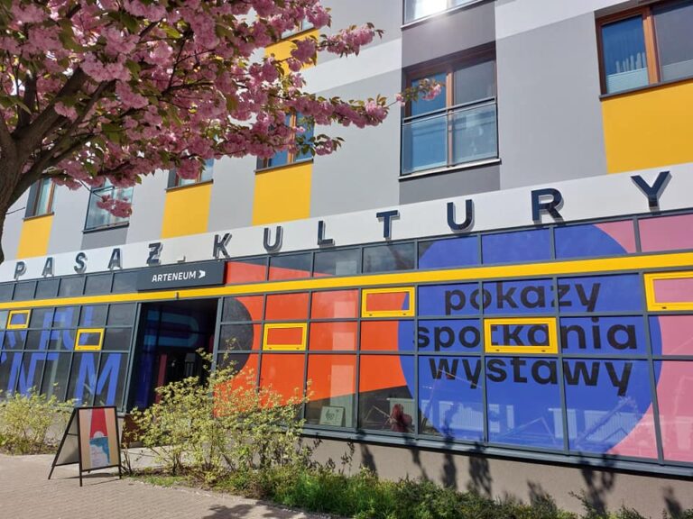 Kolorowa elewacja budynku "Pasaż Kultury" z napisem "ARTENEUM" nad wejściem, dużymi witrynami zdobionymi geometrycznymi wzorami w odcieniach pomarańczu, niebieskiego i różu oraz napisami "pokazy, spotkania, wystawy". Przed wejściem stoi tablica reklamowa, a nad budynkiem widoczne kwitnące różowe drzewo wiśni.