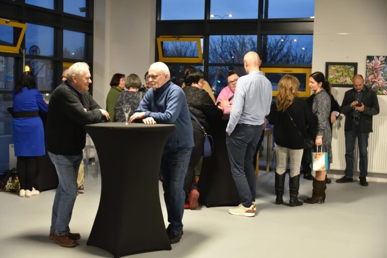 Grupa ludzi w różnym wieku rozmawiających i integrujących się podczas wieczornego spotkania towarzyskiego wewnątrz galerii, stojących przy wysokich koktajlowych stolikach nakrytych czarnym obrusem. W tle na ścianach wiszą obrazy przedstawiające kwiaty, a przez okna widać zmierzchające niebo.