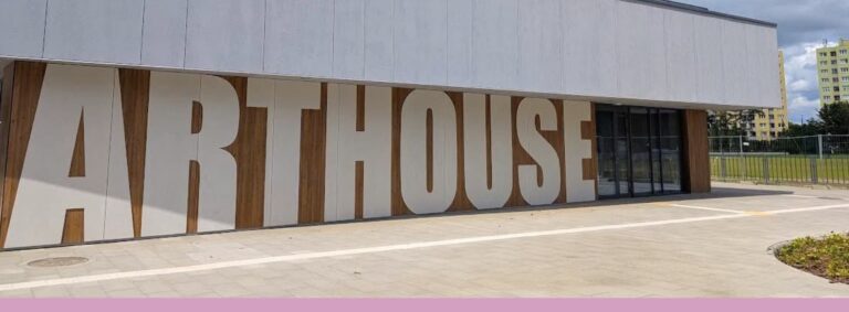 Elewacja budynku Arthouse Kotłowa – nowoczesna fasada z wielkimi literami „ARTHOUSE" wyciętymi w jasnych panelach na tle drewnianego wykończenia ściany. Przy wejściu szklane drzwi przesuwne, przed budynkiem przestronny plac z kostki.