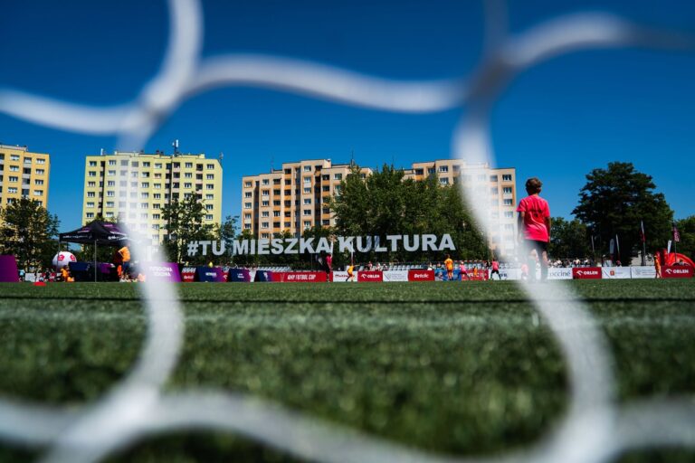 Boisko sportowe widziane przez otwory siatki bramki, w tle chłopiec w różowej koszulce, hala namiotowa oraz duży napis "#TU MIESZKA KULTURA" na trybunach. Za boiskiem widoczne wielopiętrowe bloki mieszkalne i drzewa na tle błękitnego nieba.