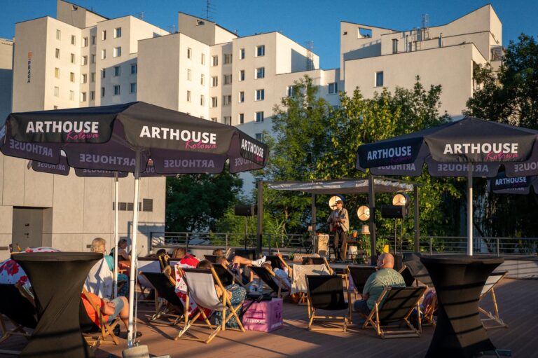 Wieczorny koncert plenerowy na tarasie Arthouse Kołowa – artysta z gitarą śpiewa na scenie oświetlonej lampami, a publiczność siedzi na leżakach pod czarnymi parasolami z logo "ARTHOUSE Kołowa". W tle białe bloki mieszkalne i drzewa.
