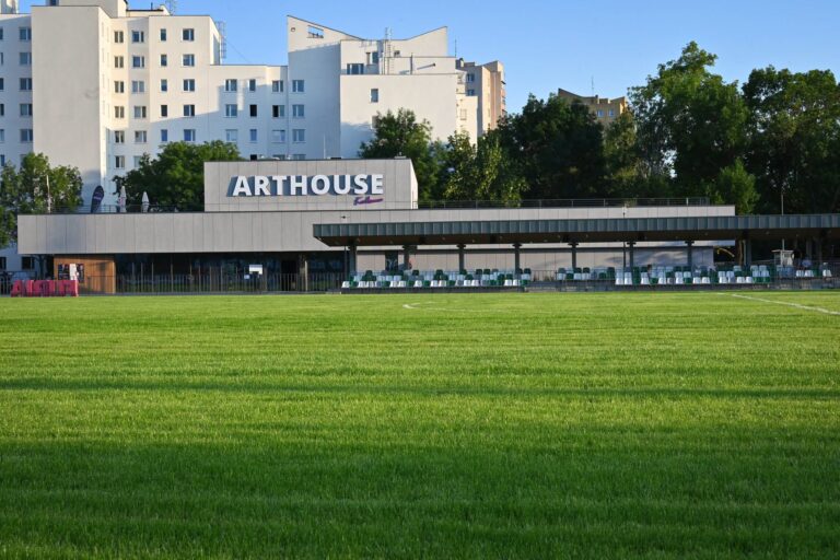 Budynek Arthouse Kołowa z dużym białym napisem "ARTHOUSE" na elewacji, widziany od strony zielonego boiska piłkarskiego z trybunami. W tle wysokie bloki mieszkalne i drzewa na tle błękitnego nieba.