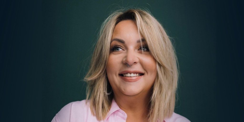 Kolorowe zdjęcie portretowe kobiety w średnim wieku. Kobieta ma blond włosy do ramion, lekko falowane, swobodnie opadające. Patrzy w górę i w bok — poza obiektyw — z szerokim, szczerym uśmiechem i rozbawioną, ciepłą miną. W nosie ma kolczyk. Ubrana jest w jasnoróżową koszulę z kołnierzykiem.