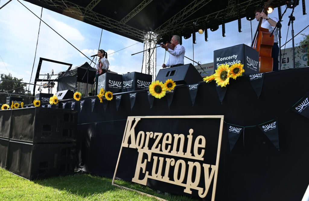 Zdjęcia z Festowalu KOrzenie Europy. Na zdjęciach widoczni są artyści na scenie, prowadzący koncert , publiczność i otoczenie koncertu - Park Bródnowski.