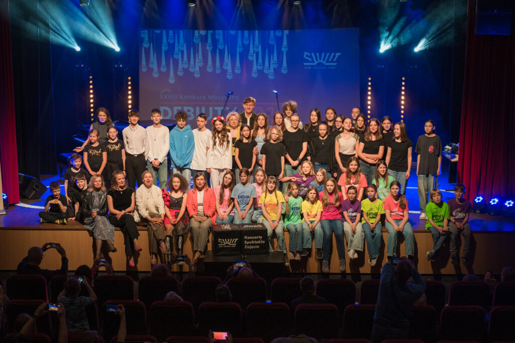 Zdjęcia z konkursu muzycznego dzieci i młodzieży Debiuty. Na o świetlonej scenie dziecięce zespołu i soliście, a przed sceną siedzi trzyosobowe jury.