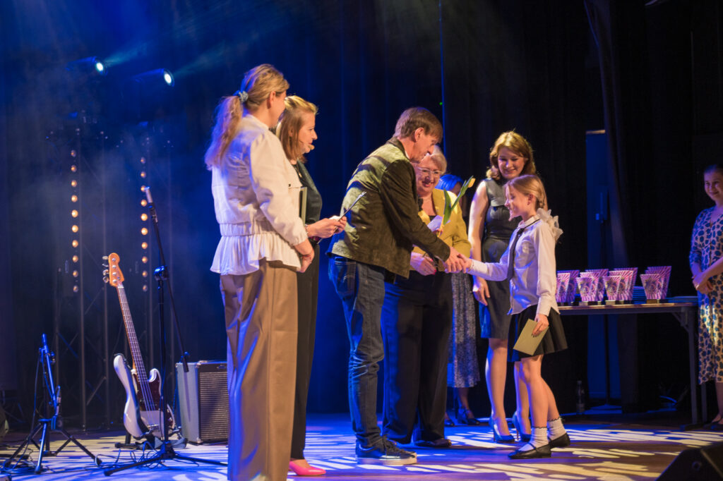 Zdjęcia z konkursu muzycznego dzieci i młodzieży Debiuty. Na o świetlonej scenie dziecięce zespołu i soliście, a przed sceną siedzi trzyosobowe jury.