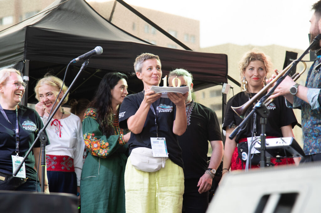 Zdjęcia z Festowalu KOrzenie Europy. Na zdjęciach widoczni są artyści na scenie, prowadzący koncert , publiczność i otoczenie koncertu - Park Bródnowski.