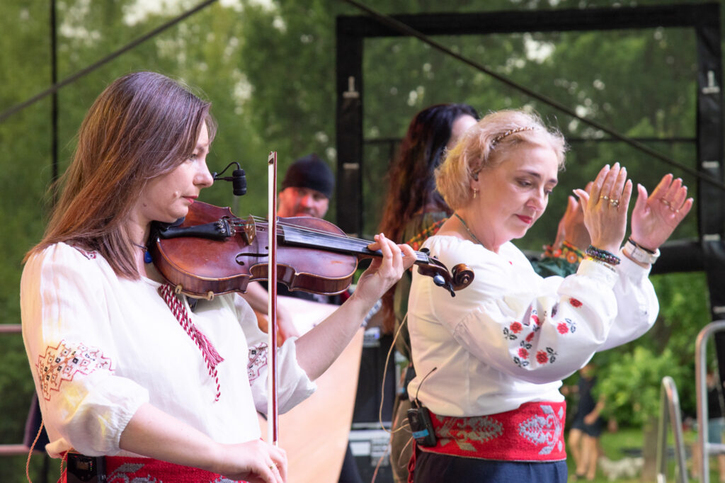Zdjęcia z Festowalu KOrzenie Europy. Na zdjęciach widoczni są artyści na scenie, prowadzący koncert , publiczność i otoczenie koncertu - Park Bródnowski.