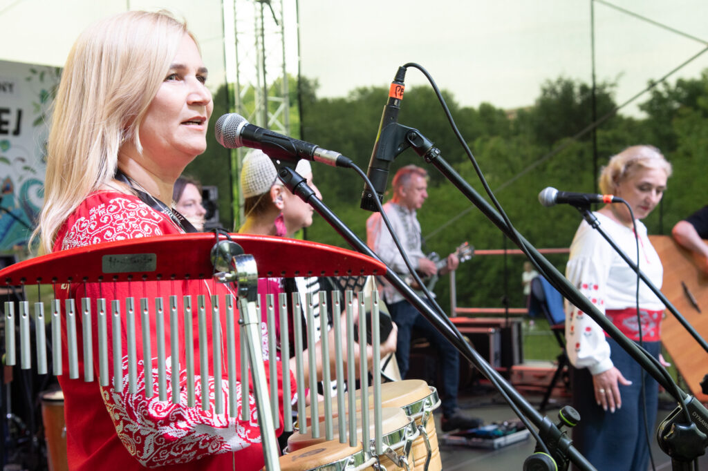 Zdjęcia z Festowalu KOrzenie Europy. Na zdjęciach widoczni są artyści na scenie, prowadzący koncert , publiczność i otoczenie koncertu - Park Bródnowski.
