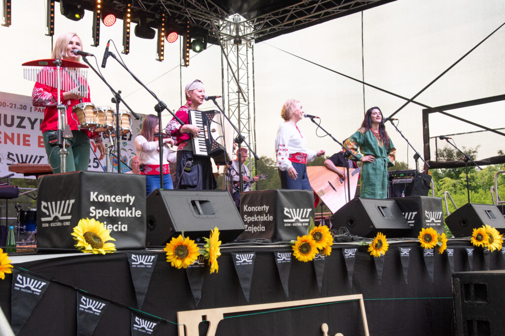 Zdjęcia z Festowalu KOrzenie Europy. Na zdjęciach widoczni są artyści na scenie, prowadzący koncert , publiczność i otoczenie koncertu - Park Bródnowski.