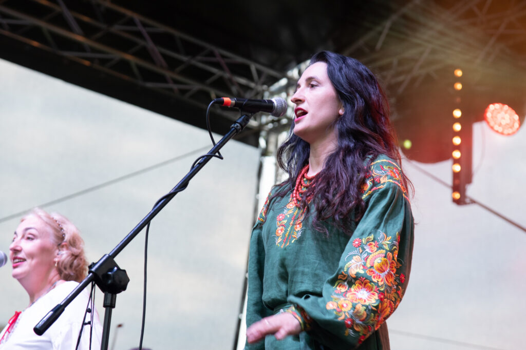 Zdjęcia z Festowalu KOrzenie Europy. Na zdjęciach widoczni są artyści na scenie, prowadzący koncert , publiczność i otoczenie koncertu - Park Bródnowski.