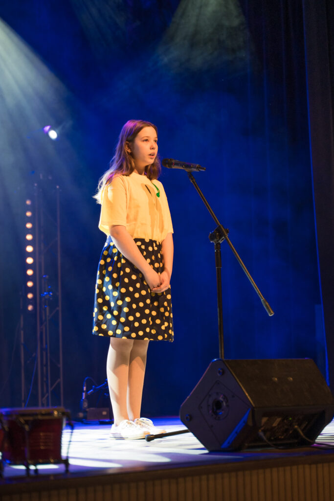 Zdjęcia z konkursu muzycznego dzieci i młodzieży Debiuty. Na o świetlonej scenie dziecięce zespołu i soliście, a przed sceną siedzi trzyosobowe jury.