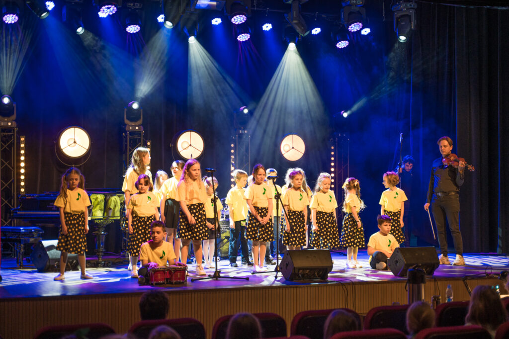 Zdjęcia z konkursu muzycznego dzieci i młodzieży Debiuty. Na o świetlonej scenie dziecięce zespołu i soliście, a przed sceną siedzi trzyosobowe jury.
