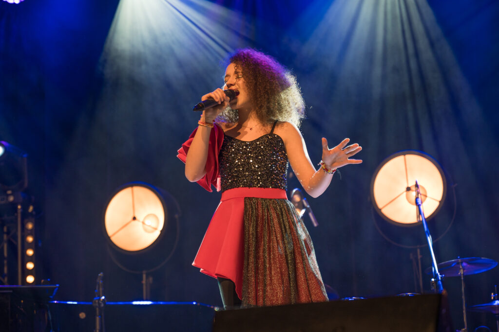 Zdjęcia z konkursu muzycznego dzieci i młodzieży Debiuty. Na o świetlonej scenie dziecięce zespołu i soliście, a przed sceną siedzi trzyosobowe jury.