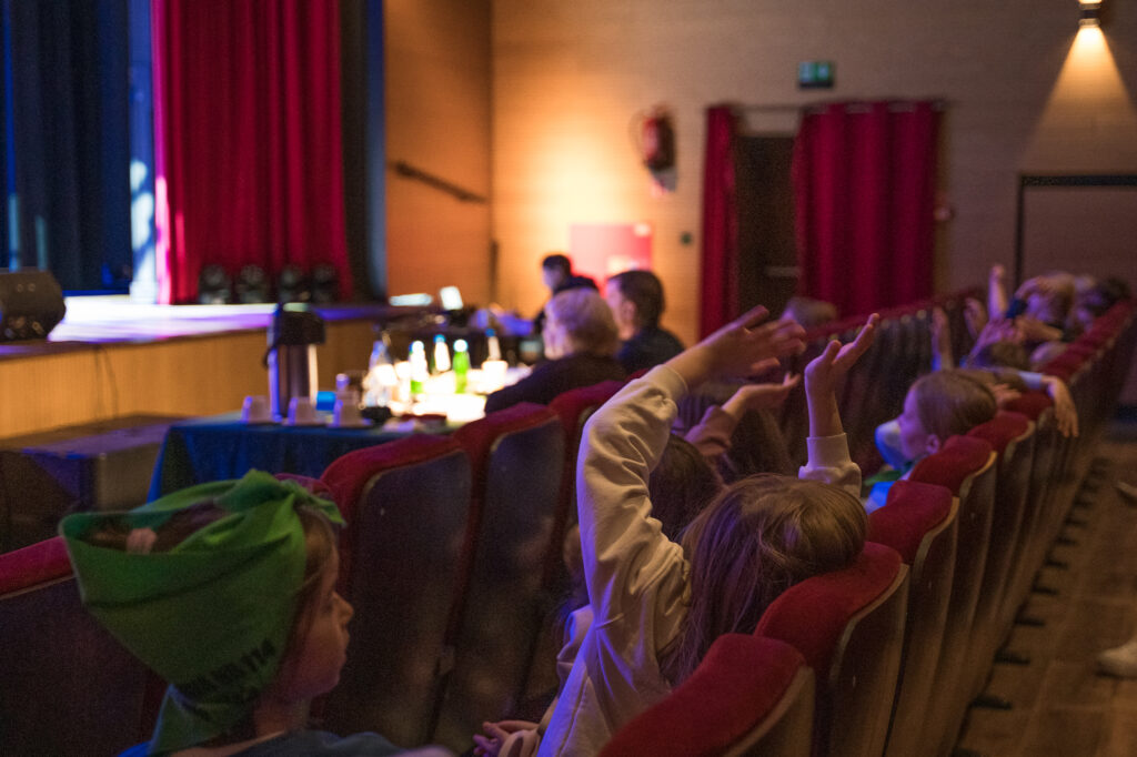 Zdjęcia z konkursu muzycznego dzieci i młodzieży Debiuty. Na o świetlonej scenie dziecięce zespołu i soliście, a przed sceną siedzi trzyosobowe jury.