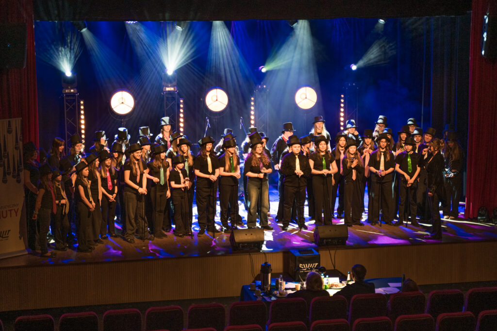 Zdjęcia z konkursu muzycznego dzieci i młodzieży Debiuty. Na o świetlonej scenie dziecięce zespołu i soliście, a przed sceną siedzi trzyosobowe jury.