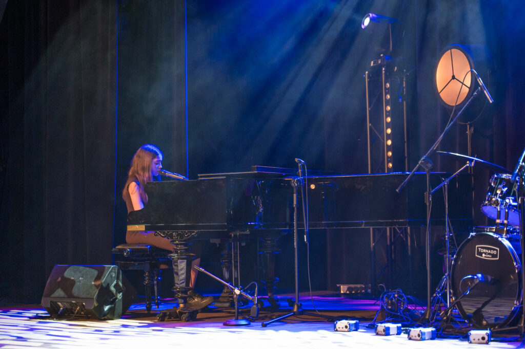 Zdjęcia z konkursu muzycznego dzieci i młodzieży Debiuty. Na o świetlonej scenie dziecięce zespołu i soliście, a przed sceną siedzi trzyosobowe jury.