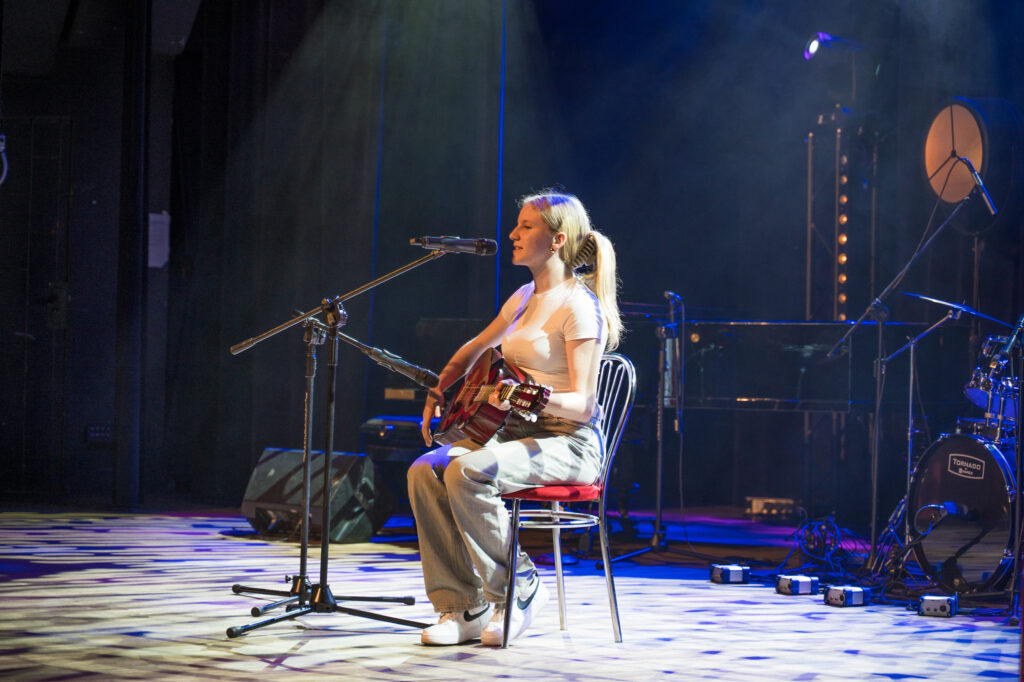 Zdjęcia z konkursu muzycznego dzieci i młodzieży Debiuty. Na o świetlonej scenie dziecięce zespołu i soliście, a przed sceną siedzi trzyosobowe jury.