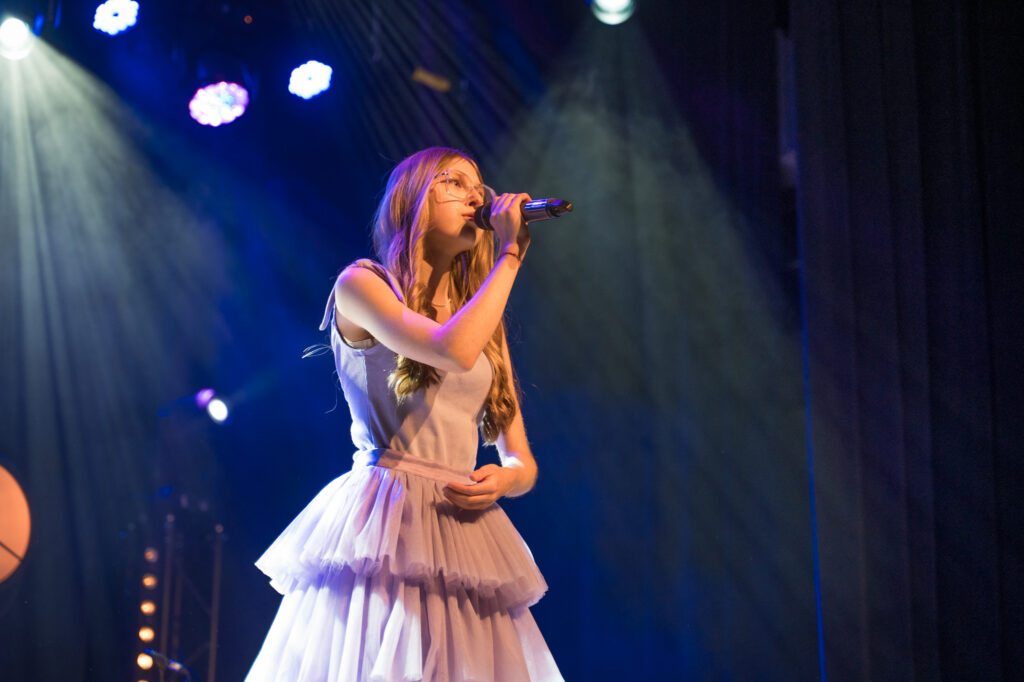 Zdjęcia z konkursu muzycznego dzieci i młodzieży Debiuty. Na o świetlonej scenie dziecięce zespołu i soliście, a przed sceną siedzi trzyosobowe jury.