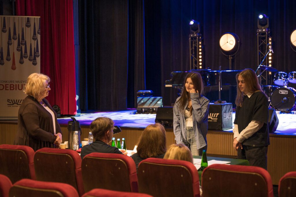 Zdjęcia z konkursu muzycznego dzieci i młodzieży Debiuty. Na o świetlonej scenie dziecięce zespołu i soliście, a przed sceną siedzi trzyosobowe jury.
