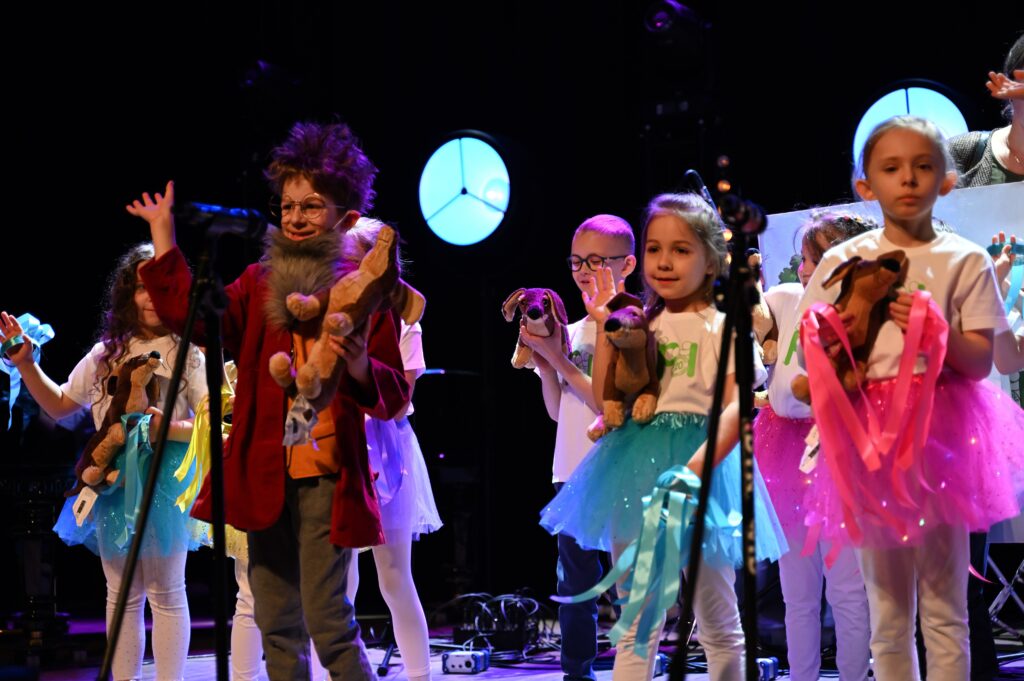 Zdjęcia z konkursu muzycznego dzieci i młodzieży Debiuty. Na o świetlonej scenie dziecięce zespołu i soliście, a przed sceną siedzi trzyosobowe jury.