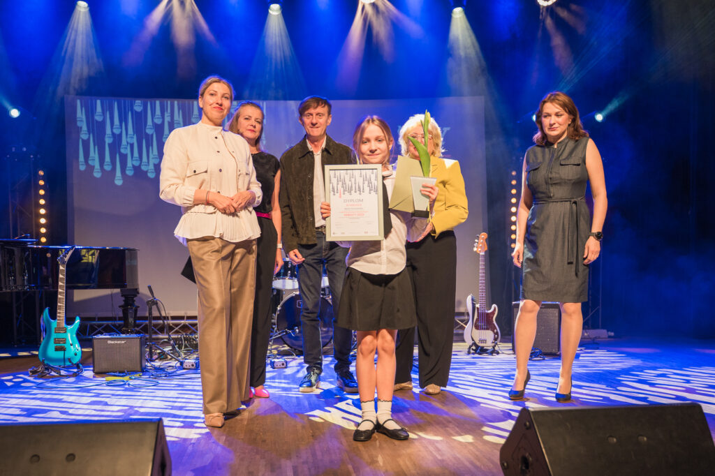 Zdjęcia z konkursu muzycznego dzieci i młodzieży Debiuty. Na o świetlonej scenie dziecięce zespołu i soliście, a przed sceną siedzi trzyosobowe jury.