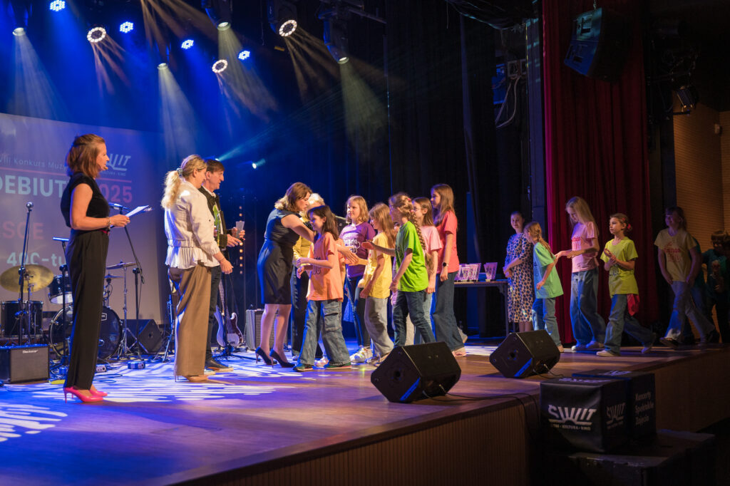 Zdjęcia z konkursu muzycznego dzieci i młodzieży Debiuty. Na o świetlonej scenie dziecięce zespołu i soliście, a przed sceną siedzi trzyosobowe jury.