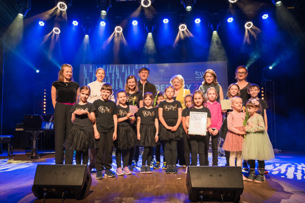 Zdjęcia z konkursu muzycznego dzieci i młodzieży Debiuty. Na o świetlonej scenie dziecięce zespołu i soliście, a przed sceną siedzi trzyosobowe jury.