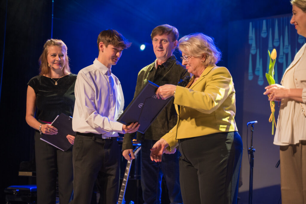 Zdjęcia z konkursu muzycznego dzieci i młodzieży Debiuty. Na o świetlonej scenie dziecięce zespołu i soliście, a przed sceną siedzi trzyosobowe jury.