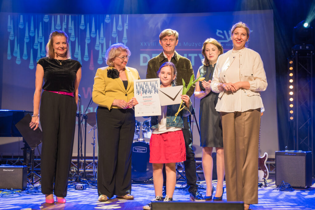 Zdjęcia z konkursu muzycznego dzieci i młodzieży Debiuty. Na o świetlonej scenie dziecięce zespołu i soliście, a przed sceną siedzi trzyosobowe jury.