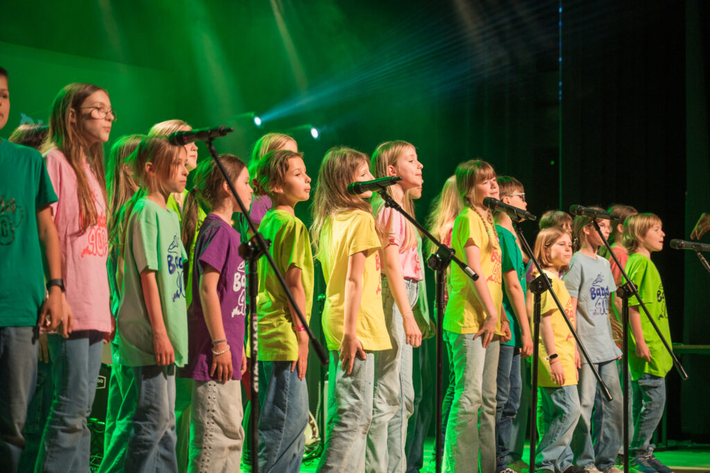 Zdjęcia z konkursu muzycznego dzieci i młodzieży Debiuty. Na o świetlonej scenie dziecięce zespołu i soliście, a przed sceną siedzi trzyosobowe jury.