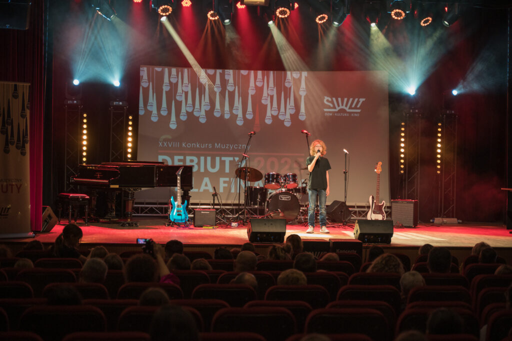 Zdjęcia z konkursu muzycznego dzieci i młodzieży Debiuty. Na o świetlonej scenie dziecięce zespołu i soliście, a przed sceną siedzi trzyosobowe jury.