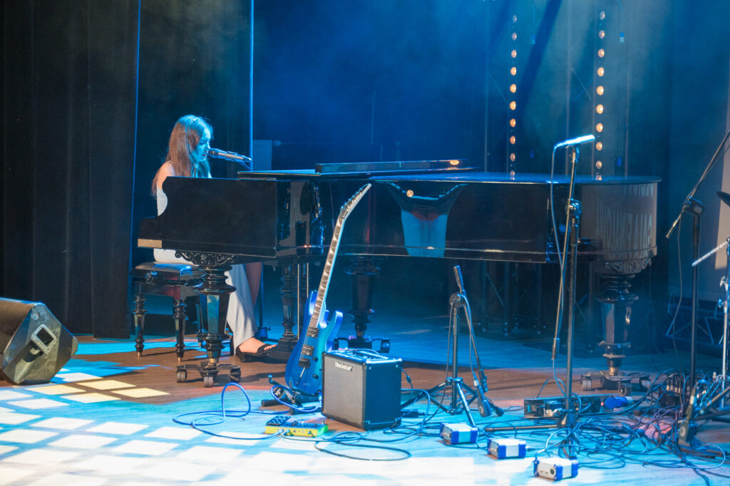 Zdjęcia z konkursu muzycznego dzieci i młodzieży Debiuty. Na o świetlonej scenie dziecięce zespołu i soliście, a przed sceną siedzi trzyosobowe jury.