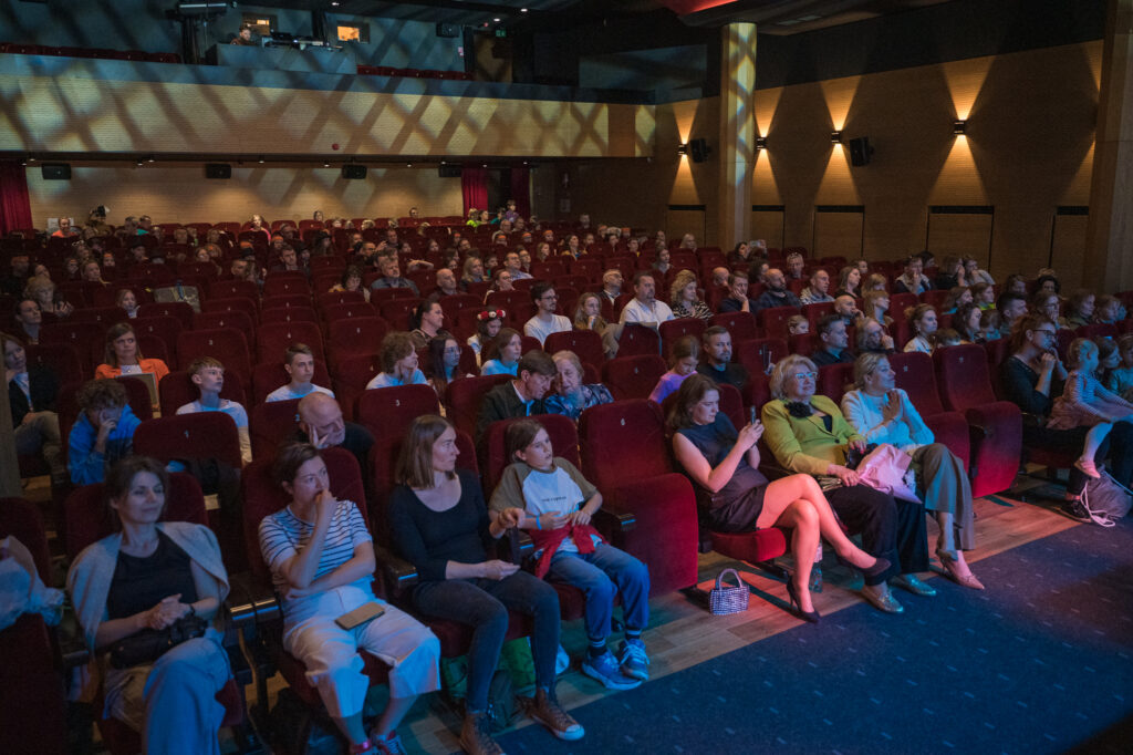 Zdjęcia z konkursu muzycznego dzieci i młodzieży Debiuty. Na o świetlonej scenie dziecięce zespołu i soliście, a przed sceną siedzi trzyosobowe jury.