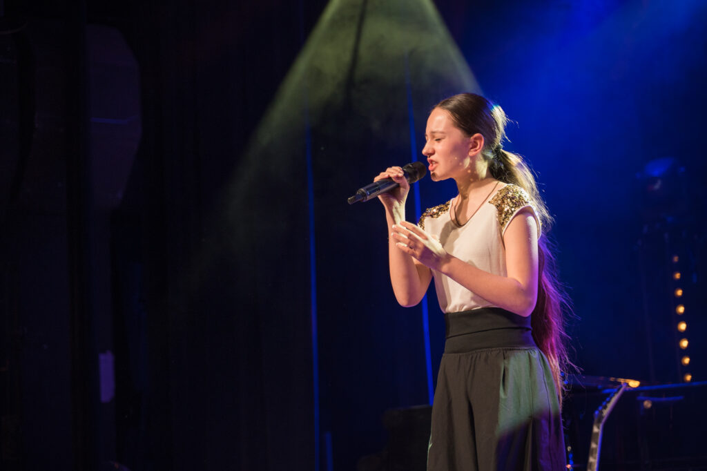 Zdjęcia z konkursu muzycznego dzieci i młodzieży Debiuty. Na o świetlonej scenie dziecięce zespołu i soliście, a przed sceną siedzi trzyosobowe jury.