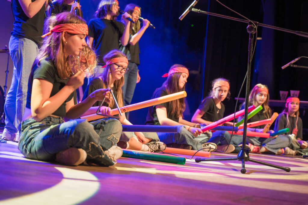 Zdjęcia z konkursu muzycznego dzieci i młodzieży Debiuty. Na o świetlonej scenie dziecięce zespołu i soliście, a przed sceną siedzi trzyosobowe jury.