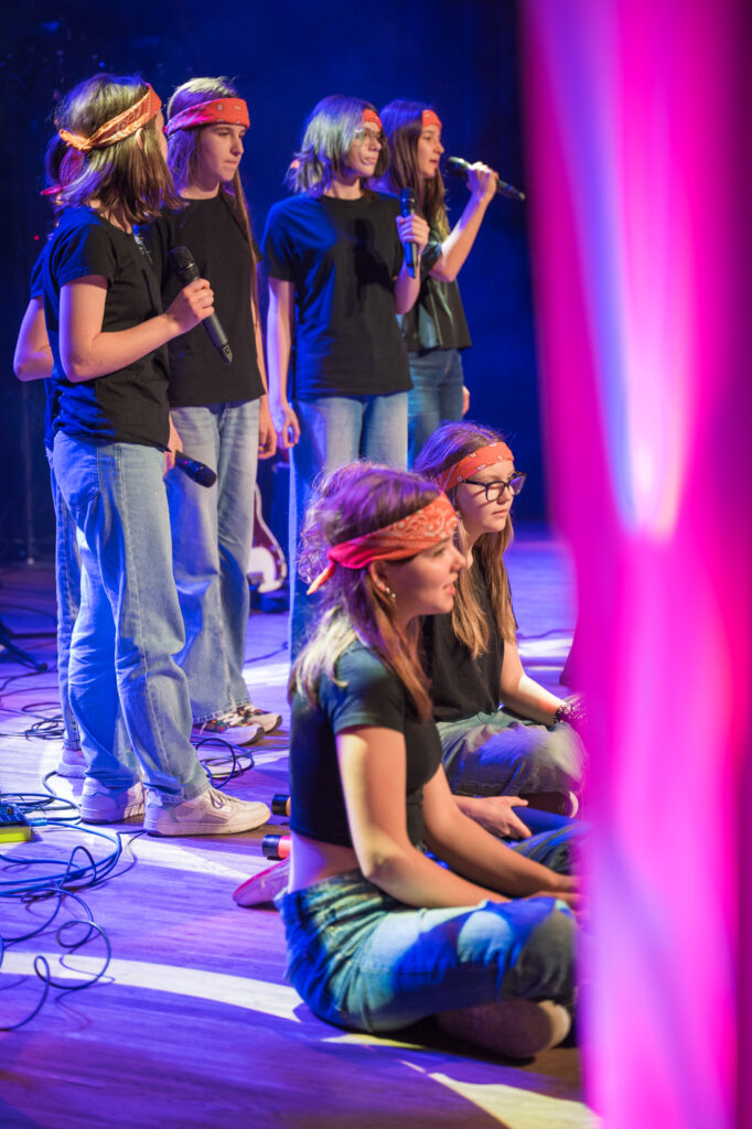 Zdjęcia z konkursu muzycznego dzieci i młodzieży Debiuty. Na o świetlonej scenie dziecięce zespołu i soliście, a przed sceną siedzi trzyosobowe jury.