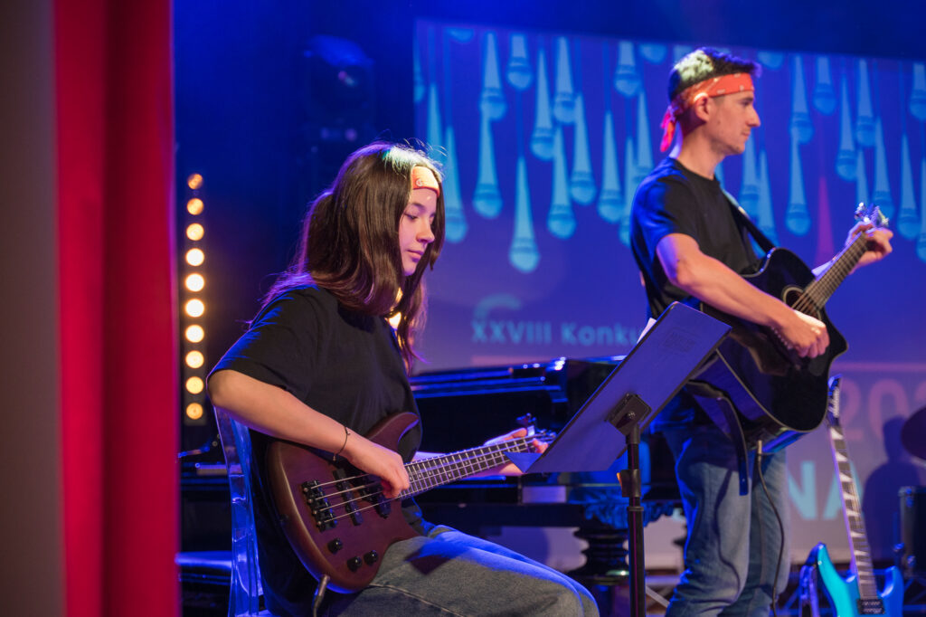 Zdjęcia z konkursu muzycznego dzieci i młodzieży Debiuty. Na o świetlonej scenie dziecięce zespołu i soliście, a przed sceną siedzi trzyosobowe jury.