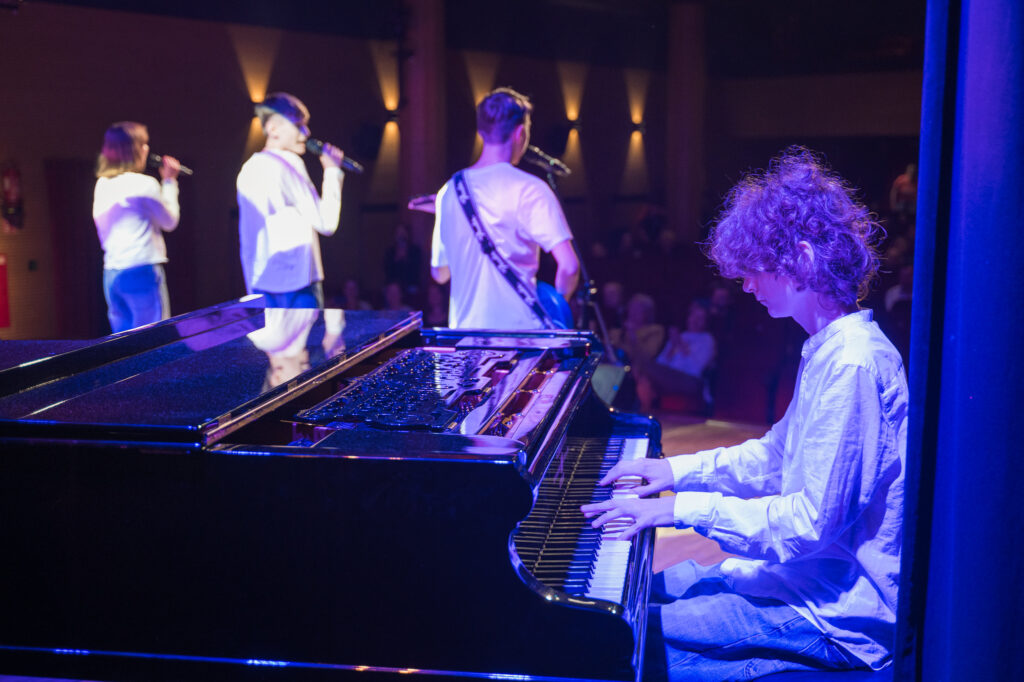 Zdjęcia z konkursu muzycznego dzieci i młodzieży Debiuty. Na o świetlonej scenie dziecięce zespołu i soliście, a przed sceną siedzi trzyosobowe jury.