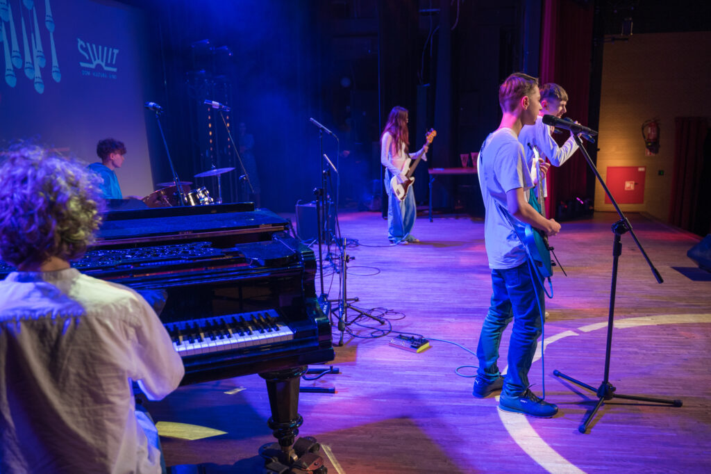 Zdjęcia z konkursu muzycznego dzieci i młodzieży Debiuty. Na o świetlonej scenie dziecięce zespołu i soliście, a przed sceną siedzi trzyosobowe jury.