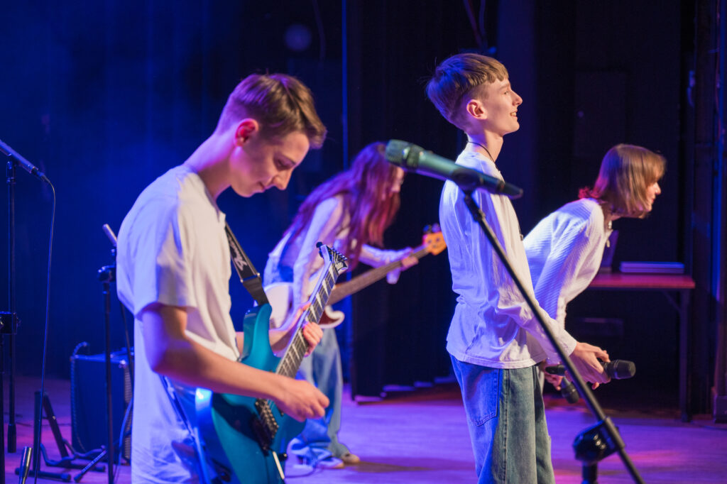 Zdjęcia z konkursu muzycznego dzieci i młodzieży Debiuty. Na o świetlonej scenie dziecięce zespołu i soliście, a przed sceną siedzi trzyosobowe jury.