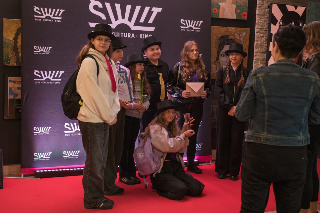 Zdjęcia z konkursu muzycznego dzieci i młodzieży Debiuty. Na o świetlonej scenie dziecięce zespołu i soliście, a przed sceną siedzi trzyosobowe jury.