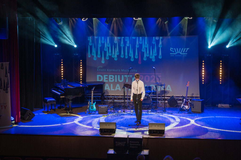 Zdjęcia z konkursu muzycznego dzieci i młodzieży Debiuty. Na o świetlonej scenie dziecięce zespołu i soliście, a przed sceną siedzi trzyosobowe jury.
