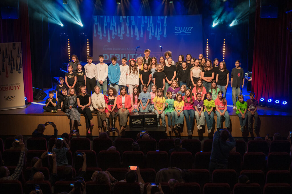 Zdjęcia z konkursu muzycznego dzieci i młodzieży Debiuty. Na o świetlonej scenie dziecięce zespołu i soliście, a przed sceną siedzi trzyosobowe jury.