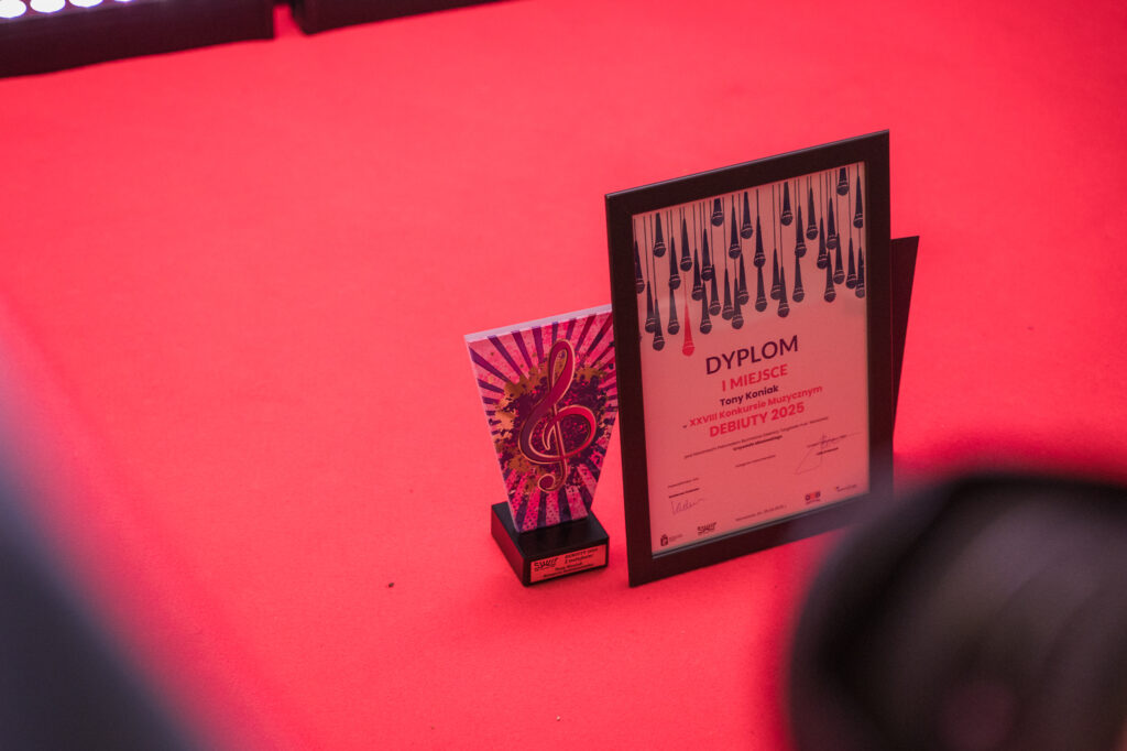 Zdjęcia z konkursu muzycznego dzieci i młodzieży Debiuty. Na o świetlonej scenie dziecięce zespołu i soliście, a przed sceną siedzi trzyosobowe jury.