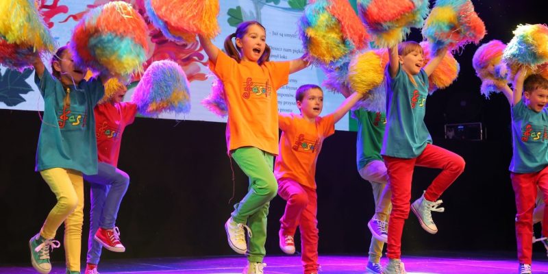 Grupa dzieci tańczy energicznie na scenie podczas występu, trzymając w uniesionych rękach kolorowe pomponiki w żółto-pomarańczowo-niebieskich barwach. Dzieci ubrane są w jednolite koszulki z logo „Fasolki" w kolorach pomarańczowym i turkusowym oraz kolorowe spodnie.
