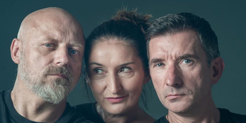 Zbliżenie twarzy trojga dorosłych ludzi na ciemnoszarym tle. Po lewej łysy mężczyzna z siwą brodą, pośrodku kobieta z ciemnymi włosami spiętymi w kok, po prawej mężczyzna z włosami zaczesanymi do tyłu. Wszyscy ubrani na czarno.