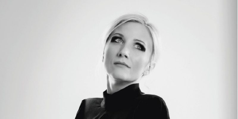 Czarno-białe zdjęcie portretowe utrzymane w minimalistycznej, eleganckiej estetyce. Wzrok kobiety skierowany ponad obiektyw, w górę, z zadumą, marzycielskim wyrazem twarzy. Kobieta ma jasne włosy zaczesane gładko do tyłu, wyraźnie zarysowane oczy podkreślone intensywnym, ciemnym makijażem. Rysy twarzy delikatne, wyraz spokojny i elegancki.