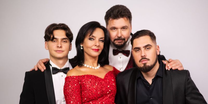 Cztery osoby pozują do oficjalnego zdjęcia na jasnym tle. Od lewej: młody mężczyzna w czarnym smokingu z czarną muszką; kobieta w czerwonej, cekinowej sukni bez ramion z perłowym naszyjnikiem i czerwonymi ustami; mężczyzna w białej koszuli z bordową muszką stojący nieco z tyłu; oraz młody mężczyzna z brodą w czarnej marynarce. Wszyscy obejmują się ramionami i patrzą w obiektyw.